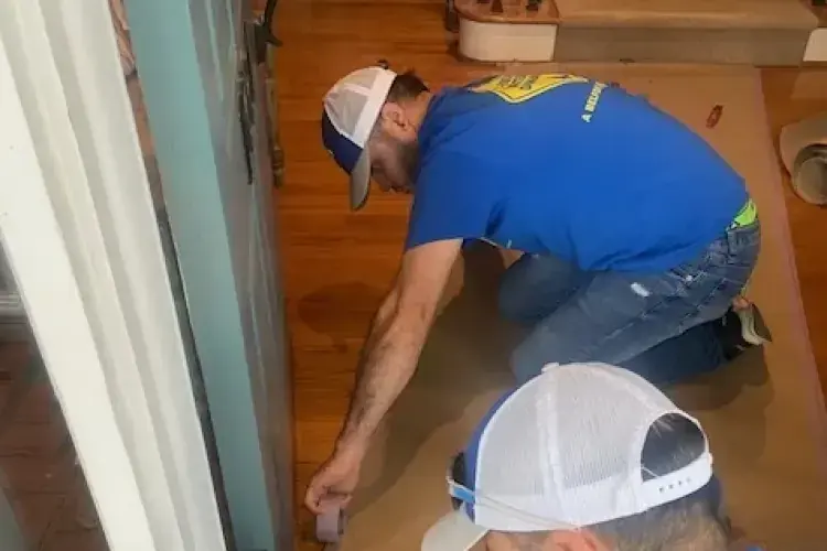 Two workers kneeling on a wood floor checking water damage at the baseboard of a Hanahan room