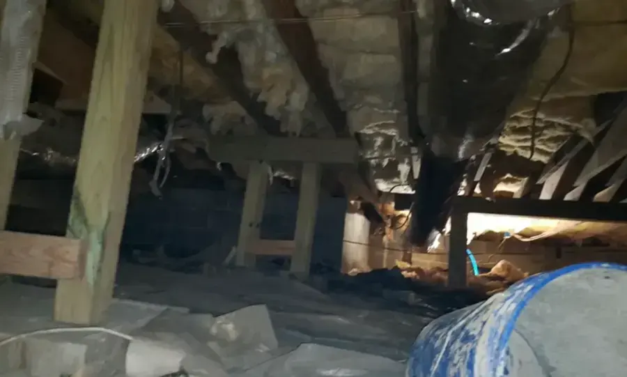 Low crawl space under a raised house with damp ground, joists and a torn ground cover, crawl space water damage in Goose Creek